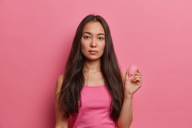 Protection menstruelle, gynécologie et concept d'hygiène féminine. Femme ethnique brune sérieuse tient la coupe menstruelle, utilise un produit absorbant zéro déchet dans la période sanguine, isolé sur un mur rose