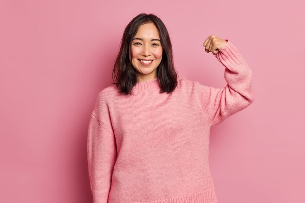 Une puissante femme brune joyeuse lève le bras et montre ses muscles démontre sa force semble confiante à la caméra sourit doucement porte un pull décontracté à manches longues