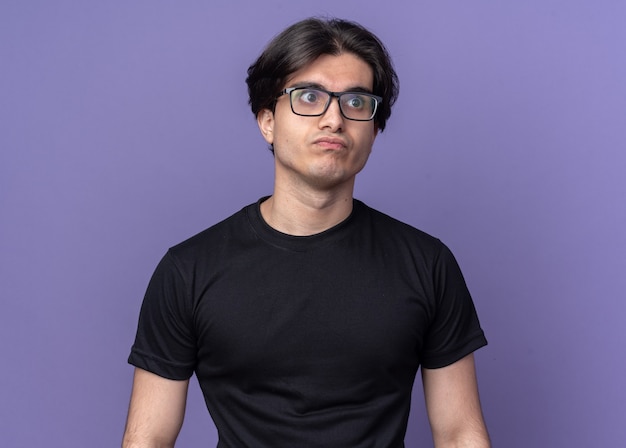 Pursing lèvres jeune beau mec portant un t-shirt noir et des lunettes isolés sur un mur violet
