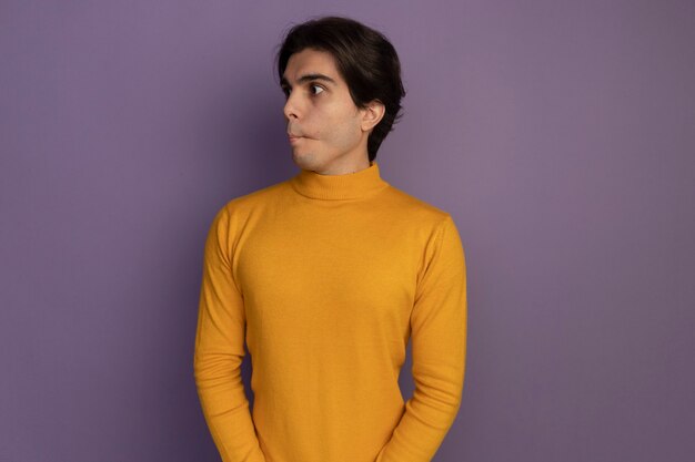 Pursing lèvres regardant côté jeune beau mec portant un pull à col roulé jaune isolé sur un mur violet avec copie espace