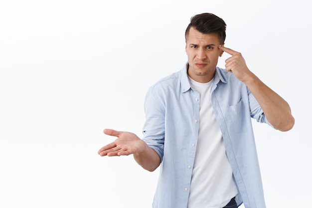 Qu'est ce qui ne va pas avec toi. Portrait d'un homme frustré et agacé tenant le doigt contre la tête et ayant l'air perplexe, levant la main avec consternation, grondant une personne folle, agissant de manière étrange ou stupide, mur blanc