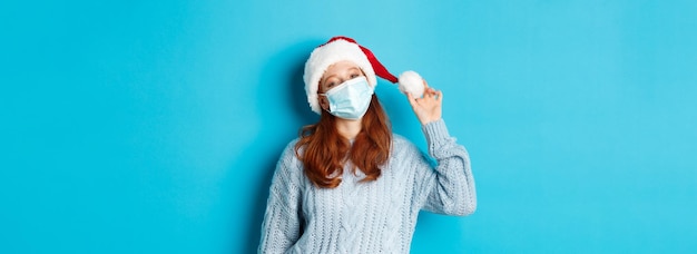 Photo gratuite quarantaine de noël et concept covid joyeuse adolescente rousse en bonnet de noel et masque facial regardant fixement