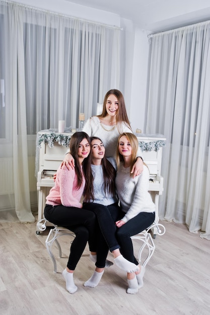 Quatre amies mignonnes que les filles portent sur des chandails chauds et un pantalon noir contre un vieux piano avec une décoration de noël à la salle blanche