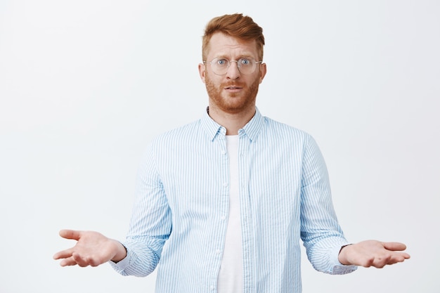 Photo gratuite ce que j'ai mal fait. confus et sombre beau mec rousse avec une barbe dans des lunettes et une chemise, debout avec une expression interrogée et étalant les paumes dans une pose désemparée, ignorant et incertain de ce qui s'est passé