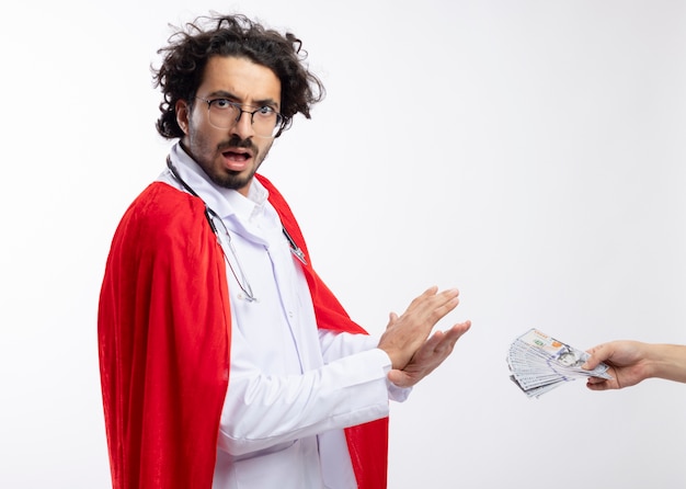 Quelqu'un remet de l'argent à un jeune homme de race blanche anxieux dans des lunettes optiques portant l'uniforme du médecin avec une cape rouge et avec un stéthoscope autour du cou faisant des gestes non avec les mains