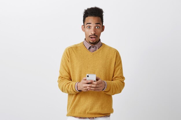 Quelqu'un a tenté de pirater son téléphone. Choqué bel homme avec une coiffure afro dans des vêtements à la mode tenant un smartphone, regardant avec une expression étonnée, surpris sur un mur gris