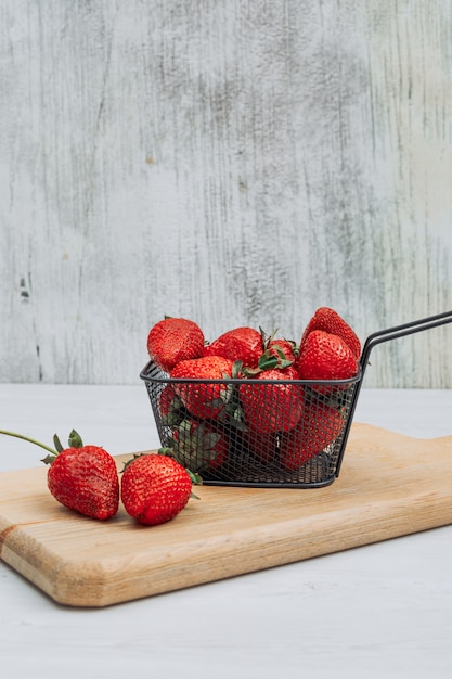 Photo gratuite quelques fraises avec plusieurs fraises autour dans un panier noir sur une planche à découper en bois et fond blanc, vue latérale