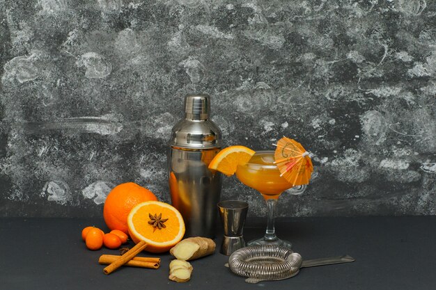 Quelques oranges avec du jus dans un verre, thermos, gingembre et tranches sur une surface texturée, vue latérale.