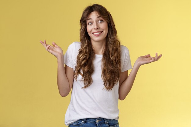 Qui s'en soucie, amusez-vous. Insouciante indifférente sortante heureuse jeune femme hausse les mains sur le côté désemparé indifférent apathique au sujet indifférent souriant largement insouciant fond jaune