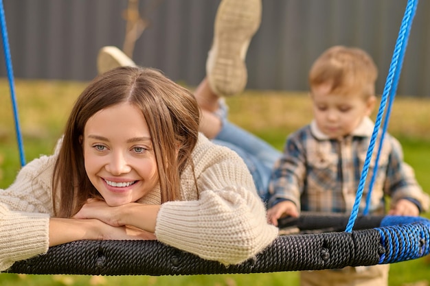 Équilibre intérieur. Belle jeune femme souriante aux longs cheveux blonds en pull et jeans allongé sur la balançoire à l'air heureux à côté et enfant debout le beau jour