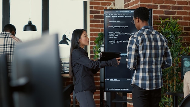 Équipe de codeurs analysant le code sur un écran de télévision mural comparant les erreurs à l'aide d'une tablette numérique à côté d'un programmeur travaillant avec un réseau de neurones. Ingénieurs logiciels collaborant sur un projet de groupe de codage.