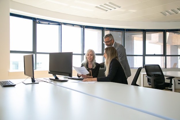 Équipe commerciale positive discutant du rapport, assis à la table de réunion avec moniteur, tenant à la recherche de documents. Réunion d'affaires ou concept de travail d'équipe