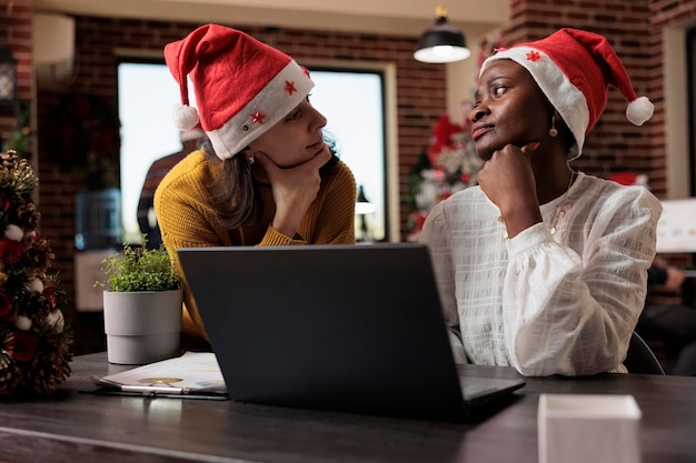 Équipe diversifiée de collègues avec un bonnet de noel travaillant sur un ordinateur portable, assis dans un bureau festif rempli de décorations et d'ornements de noël. Faire du travail d'équipe et célébrer les vacances.