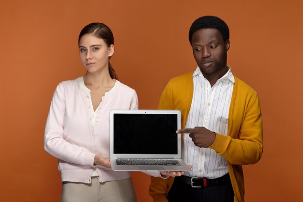 Équipe interraciale élégante de deux collègues mand noir et femme blanche bénéficiant d'une connexion Internet sans fil tout en utilisant un ordinateur portable avec un écran noir vierge avec copyspace pour votre information
