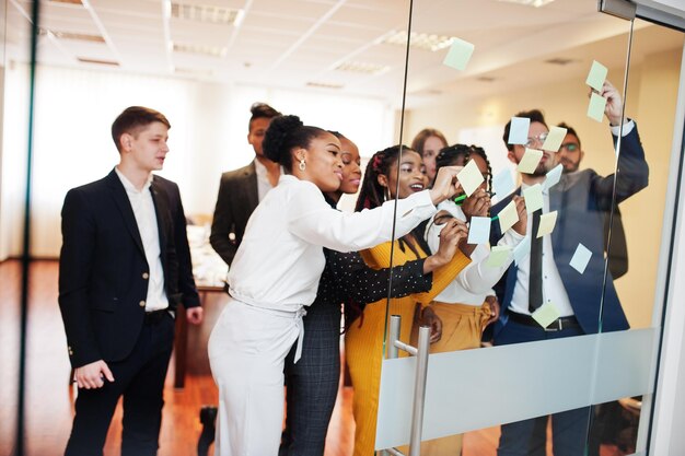 Équipe de jeunes multiculturels pointant sur du verre avec des notes en papier colorées Groupe diversifié d'employés masculins et féminins en tenue de soirée à l'aide d'autocollants