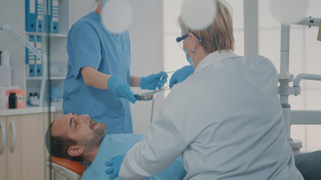 Équipe de spécialistes effectuant une intervention chirurgicale sur un homme souffrant de maux de dents, utilisant des outils dentaires et un instrument pour l'opération de stomatologie. Dentiste et infirmière extrayant des dents à la clinique de soins bucco-dentaires.