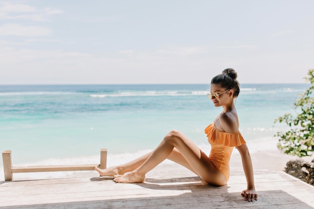 Raffiné Mince Jeune Femme Posant Sur La Plage. Agréable Fille Bronzée En Lunettes De Soleil Et Maillot De Bain élégant Se Détendre Sur La Côte De L'océan Avec Un Sourire Timide.