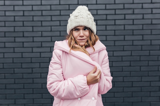 Raffinée jeune femme en tenue d'hiver faisant des grimaces. Tir extérieur d'une fille caucasienne insouciante en manteau rose et bonnet tricoté.