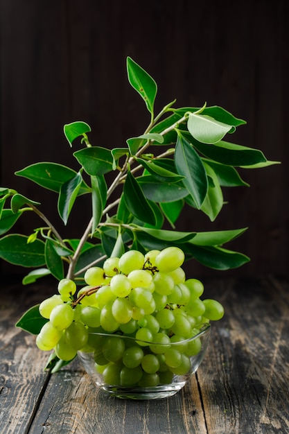 Raisins avec des feuilles sur une branche dans un bol en verre sur une surface en bois, vue latérale.