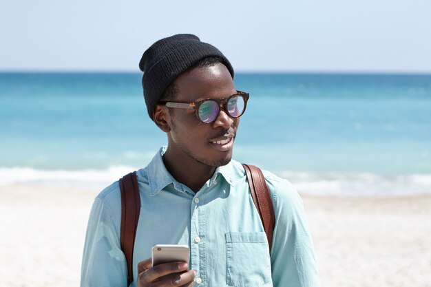 Randonneur afro-américain fatigué en chapeau et lunettes utilisant l'application de service de taxi en ligne sur téléphone portable pour demander un taxi tout en ayant soif, à la recherche d'un endroit pour prendre une boisson fraîche