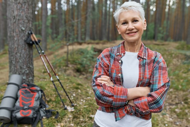 Randonneur femme d'âge moyen attrayant énergique posant à l'extérieur, choisissant un mode de vie actif, voyageant seul avec un sac à dos et des bâtons nordiques pour marcher, garder les bras croisés et souriant à la caméra