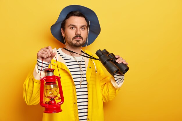 Randonneur masculin sérieux porte une lampe à gaz, utilise des jumelles lors d'un voyage de randonnée, vêtu d'un imperméable, regarde avec confiance la caméra isolée sur un mur jaune