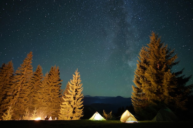 Randonneurs se reposant à côté d'un feu de joie lumineux près de tentes touristiques illuminées sur un terrain de camping dans des bois de montagne sombres sous un ciel nocturne avec des étoiles scintillantes. mode de vie actif et concept de vie en plein air.