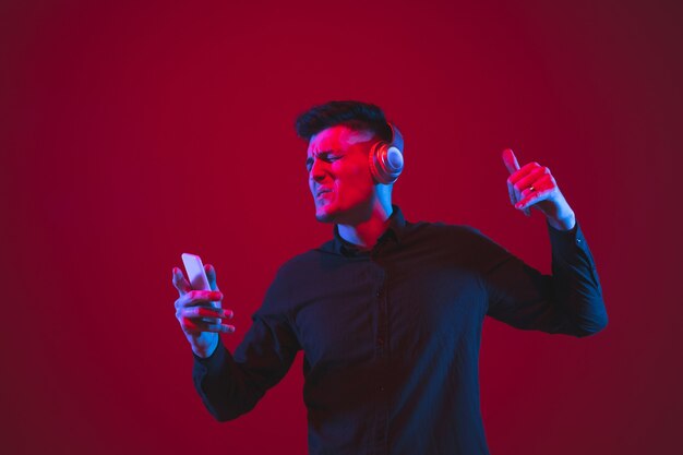 Ravi d'écouter de la musique. Portrait d'un jeune homme caucasien isolé sur un mur rouge à la lumière du néon. Beau modèle masculin. Concept d'émotions humaines, d'expression faciale, de culture des jeunes.