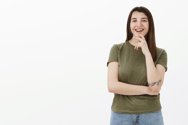 Ravi Et Heureux Jeune étudiante Ambitieuse Avec Tatouage Et Cheveux Bruns Touchant Le Menton En Riant Et Souriant Heureux Comme L'idée D'aimer, Le Plan De Réflexion Est Excellent Debout Satisfait Sur Le Mur Gris