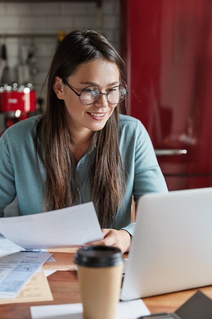 Ravie et heureuse jeune femme crée un plan réussi pour les opérations bancaires, détient des documents papier, regarde positivement l'ordinateur portable, s'assoit à la table de la cuisine.