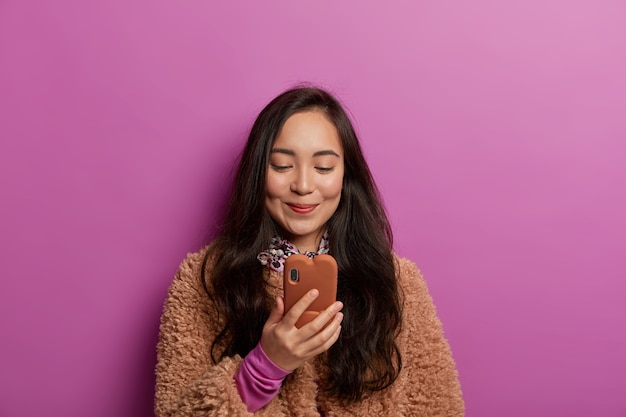 Ravie de jolie jeune femme asiatique aux cheveux noirs, lit des nouvelles intéressantes, a une dépendance aux gadgets, ravie de recevoir un message, vêtue d'un manteau brun