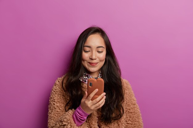 Ravie de jolie jeune femme asiatique aux cheveux noirs, lit des nouvelles intéressantes, a une dépendance aux gadgets, ravie de recevoir un message, vêtue d'un manteau brun