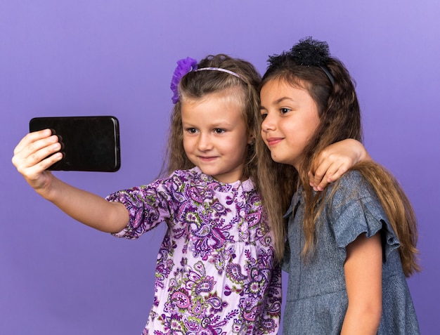 Ravies De Petites Jolies Filles Regardant Un Téléphone Prenant Un Selfie Isolé Sur Un Mur Violet Avec Un Espace De Copie