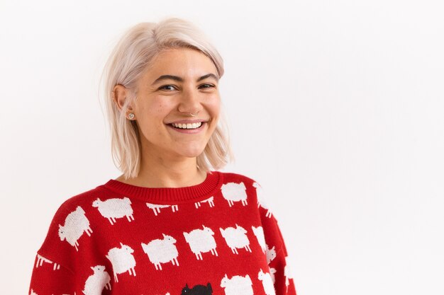 Réactions et sentiments humains. Portrait d'adorable jolie fille avec anneau dans le nez posant dans un pull rouge élégant avec des agneaux au mur blanc vierge, avec un sourire rayonnant heureux