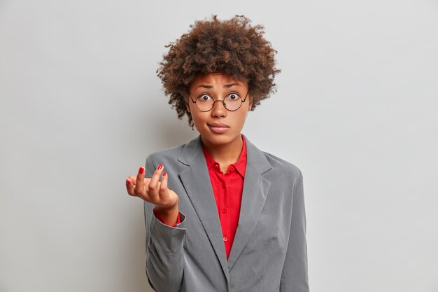 Une réalisatrice confuse et perplexe, propriétaire d'une société commerciale, hausse les épaules avec perplexité, habillée formellement, porte des lunettes rondes