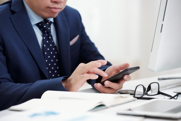 Recadré SMS d'homme d'affaires sur son téléphone assis au bureau avec ses lunettes et un ordinateur portable sur le bureau