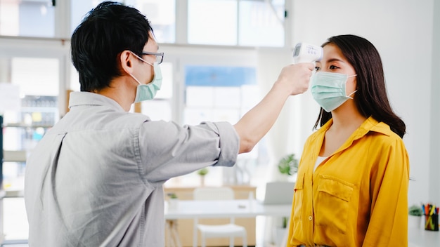 La réceptionniste masculine d'Asie portant un masque protecteur utilise un thermomètre infrarouge ou un pistolet thermique sur le front du client avant d'entrer dans le bureau. Mode de vie nouveau normal après le virus corona.