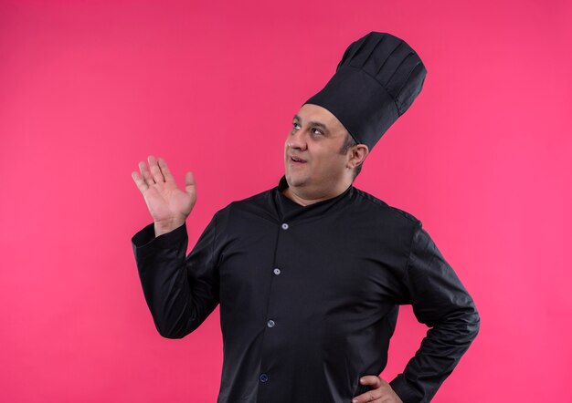 À la recherche à côté homme d'âge moyen cuisinier en uniforme de chef propagation part mettant la main dans la hanche sur un mur rose isolé