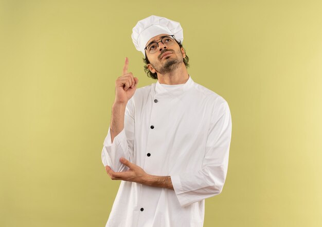 À la recherche de heureux jeune homme cuisinier portant l'uniforme de chef et des lunettes pointe vers le haut