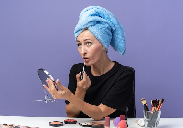 À la recherche d'une jeune belle fille assise à table avec des outils de maquillage essuyant les cheveux dans une serviette tenant un miroir appliquant un brillant à lèvres isolé sur un mur bleu