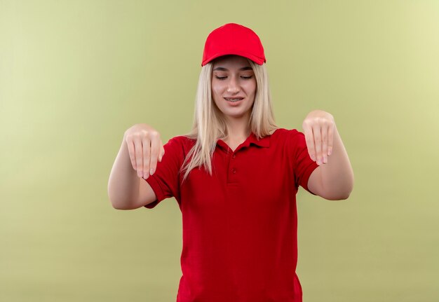 À la recherche vers le bas de livraison jeune fille portant un t-shirt rouge et une casquette en orthèse dentaire montrant prendre le geste sur fond vert isolé