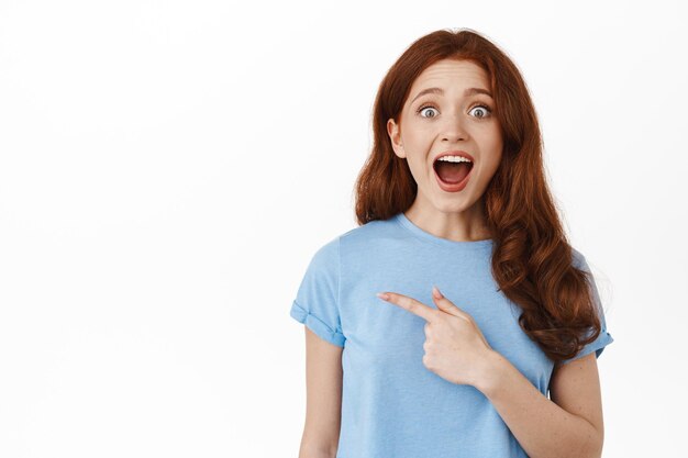 Des réductions impressionnantes. Une fille rousse étonnée halète et dit wow, vérifiant le logo promotionnel, pointant le doigt vers la gauche et réagissant à la publicité super cool, debout sur fond blanc