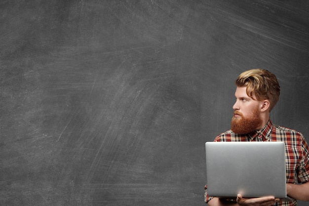 Réfléchi bel étudiant en chemise à carreaux à la mode tenant un ordinateur portable générique dans ses mains, faisant des tâches à domicile, regardant loin au tableau blanc