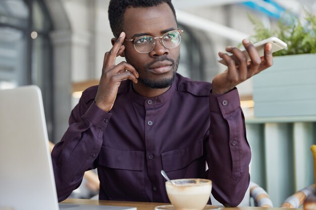 Réfléchi entrepreneur masculin à la peau sombre dans les lunettes, a une pause-café après un travail acharné, fait un rapport d'activité sur un ordinateur portable