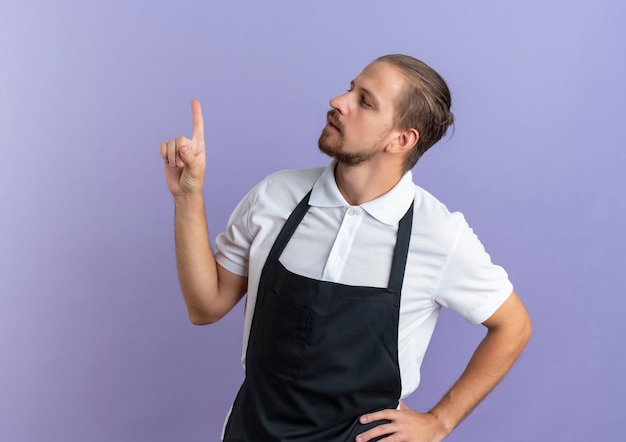 Réfléchi jeune beau coiffeur portant l'uniforme mettant la main sur la taille et levant le doigt à côté isolé sur mur violet