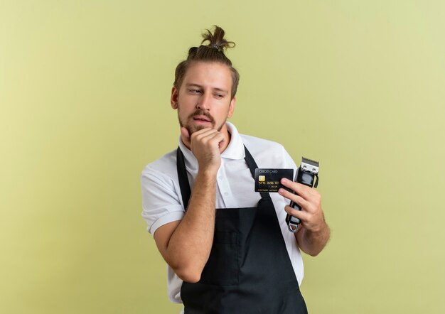 Réfléchi jeune beau coiffeur tenant une carte de crédit et une tondeuse à cheveux à côté isolé sur mur vert olive
