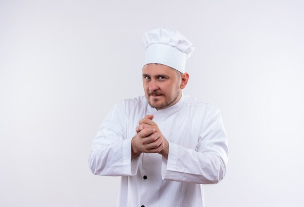 Réfléchi jeune beau cuisinier en uniforme de chef gardant les mains ensemble sur un espace blanc isolé