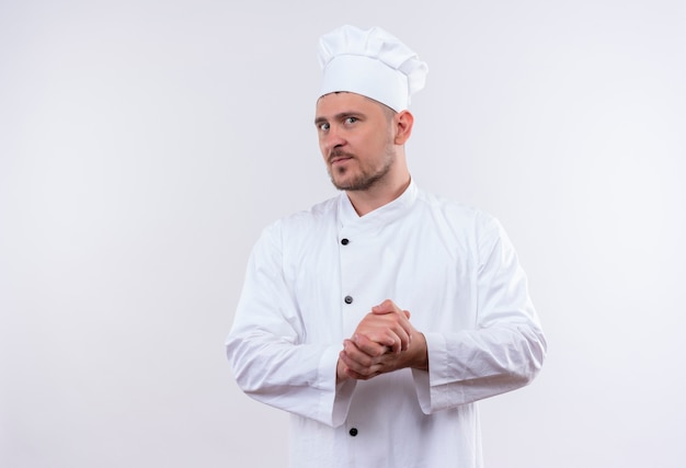 Réfléchi jeune beau cuisinier en uniforme de chef gardant les mains ensemble sur un espace blanc isolé