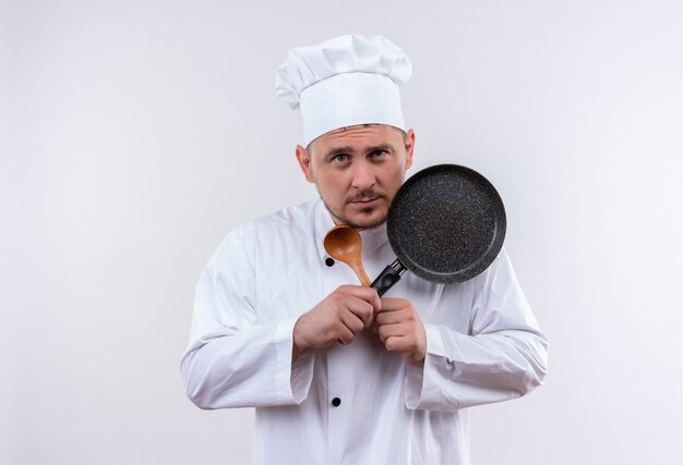 Réfléchi jeune beau cuisinier en uniforme de chef tenant cuillère et poêle à frire isolé sur espace blanc