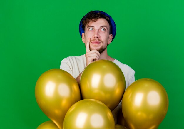 Réfléchi jeune beau mec de parti slave portant chapeau de fête debout derrière des ballons en gardant la main sur le menton à côté isolé sur fond vert